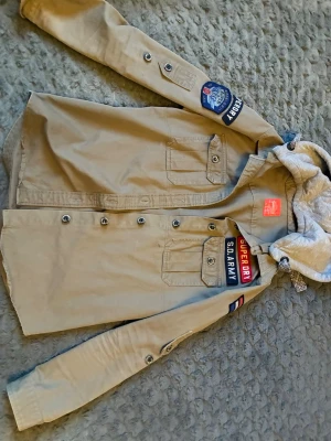 Beige overshirt med patchar Superdry - Stilren beige overshirt från Superdry med coola patchar och broderier på bröst och ärm. Har grå huva i sweatshirtmaterial, knappstängning och två bröstfickor med lock. Perfekt för lager-på-lager och streetwear-vibe.
