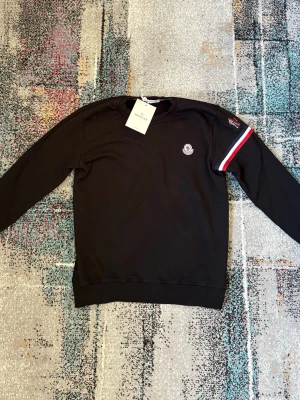 Svart Moncler sweatshirt med logga - Svart sweatshirt från Moncler med rund halsringning och Moncler-logga på bröstet. Detalj med röd, vit och blå rand på ena ärmen samt ribbade muddar vid ärmslut och nederkant. Perfekt för en stilren och sportig look.