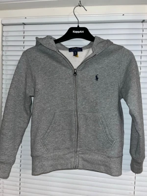 Grå hoodie från Polo Ralph Lauren - Snygg grå hoodie från Polo Ralph Lauren med dragkedja framtill och klassisk blå logga på bröstet. Hoodien har huva och två fickor framtill. Säljer denna för den är för liten, har inte använt den mycket men väldigt skön och inte smutsig. Storleken är 140-134, fick inte med det på bilden.
