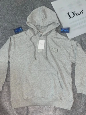 Grå hoodie från Dior med logga - Snygg grå hoodie från Dior med broderad logga på bröstet. Klassisk modell med huva och dragsko samt känguruficka framtill. Tillverkad i mjukt material som känns skönt mot huden. Perfekt för en avslappnad och trendig look.