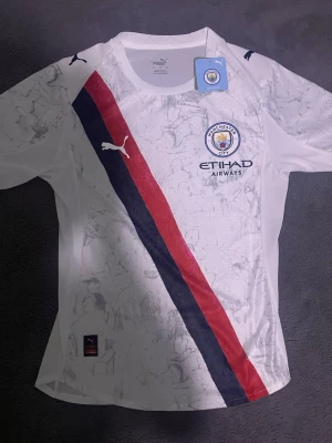 Manchester City matchtröja Puma - Säljer en vit Manchester City matchtröja från Puma med röd och marinblå diagonal rand, klubbmärke och Etihad Airways-logga på bröstet. Tröjan har korta ärmar och är tillverkad i ett lätt och ventilerande material, perfekt för fotbollsträning eller match. Helt ny oanvänd 