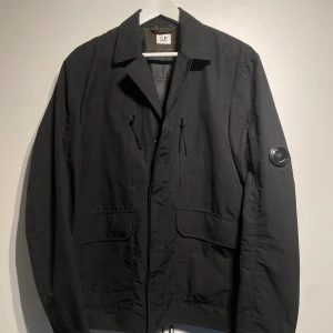 C.P. Company Overshirt  - Stilren svart jacka från C.P. Company med klassisk krage och två bröstfickor utan dragkedja. Jackan har även två stora fickor framtill och en ikonisk linsdetalj på vänster ärm. Sorlek 50/ L men passar M. Nypris 3600kr. Jackan är i bra skick och ser nästintill ny ut. Fler bilder finns i DM! 
