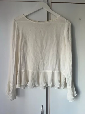 Blus med plisserad kant från Minimum  - Snygg vit blus med långa ärmar och plisserad volangkant både nedtill och vid ärmsluten. Blusen har en rak passform och rund halsringning. Perfekt för en stilren och trendig look.