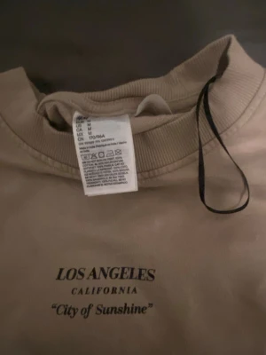 Beige sweatshirt Los Angeles print - Beige sweatshirt från H&M med trycket 'LOS ANGELES CALIFORNIA City of Sunshine' på bröstet. Tröjan har rund halsringning och långa ärmar. Tillverkad i mjuk bomullsmix, perfekt för en avslappnad streetwear-look.