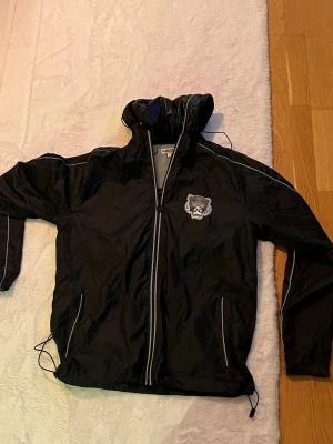 Svart vindjacka från Kenzo REFLECTIVE - Svart vindjacka från Kenzo med dragkedja, huva och vita detaljer längs ärmar och dragkedja. Jackan har Kenzo-tryck med tiger på bröstet och stort tryck på ryggen. Justerbar nederkant