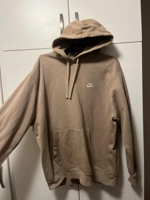 Beige hoodie från Nike med huva - Snygg beige hoodie från Nike med klassisk logga broderad i vitt på bröstet. Hoodien har en stor känguruficka framtill, ribbade muddar och justerbar dragsko i huvan. Perfekt för en avslappnad och trendig stil.