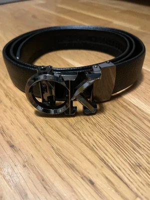 Svart Calvin Klein bälte med metallspänne - Stilrent svart bälte från Calvin Klein med ett stort, blankt metallspänne i form av CK-loggan. Bältet är gjort i skinn och har en klassisk, tidlös design som passar till många olika outfits.