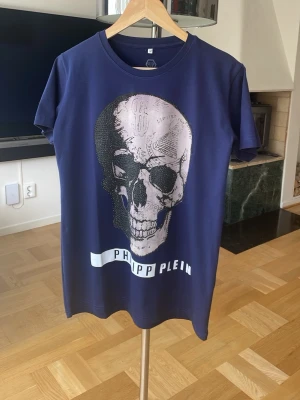 Blå t-shirt med dödskalle från Philipp Plein - Cool blå t-shirt från Philipp Plein med stort dödskalletryck framtill och märkets logga i vitt. T-shirten har rund hals och korta ärmar. Materialet känns mjukt och skönt, perfekt för en edgy streetstyle-look.