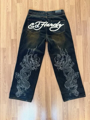 Ed Hardy jeans med drakmotiv - Säljer ett par brun svarta Ed Hardy jeans med bred passform och stora vita drakmotiv på båda benen. Bakfickorna har broderad Ed Hardy-logga och framtill finns en mindre logga. Jeansen är i denim och har en cool streetwear-vibe.