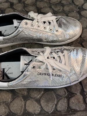 Silvriga sneakers från Calvin Klein Jeans - Snygga silvriga sneakers från Calvin Klein Jeans med reptilmönstrad yta och vita snören. Skorna har låg profil och logotypen är tryckt på sidan. Perfekt för dig som vill sticka ut med ett metallic-glansigt statement.