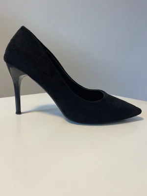 Svarta klassiska pumps i mocka - Säljer ett par svarta pumps med spetsig tå och smal hög klack. Skorna är i mockaimitation och har en elegant, tidlös design som passar till många olika looks. Perfekta för dig som vill ha en stilren och chic sko till garderoben.