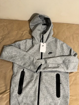 Grå Nike hoodie med dragkedja - Säljer en snygg grå hoodie från Nike med svart logga på bröstet. Hoodien har hel dragkedja, två svarta dragkedjeförsedda fickor och en stor huva. Materialet är mjukt och bekvämt, perfekt för chill eller träning. Taggen sitter kvar.