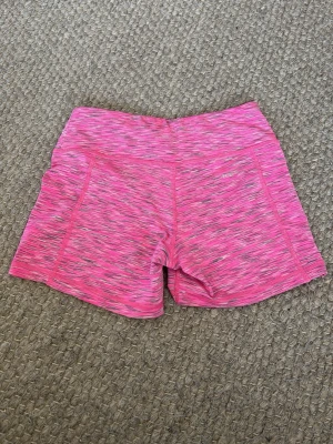 Rosa träningsshorts  - Säljer ett par rosa träningsshorts  med mönster i grått och vitt. De är högmidjade och har en tight passform, perfekta för gymmet eller löpning. Materialet är syntet och ger en skön känsla och bra rörelsefrihet.