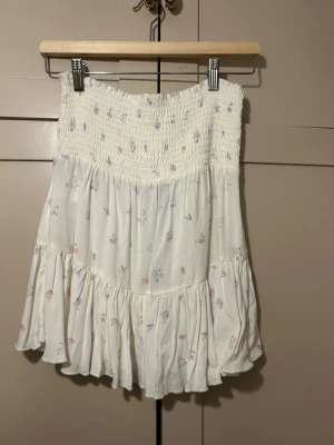 vit kortkjol med små blommor - Söt vit kortkjol med smock upptill och volang nertill. Kjolen har ett diskret blommigt mönster i pastellfärger och är gjord i ett mjukt bomullsmaterial.