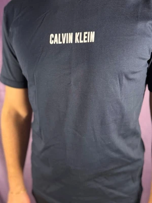 Marinblå Calvin Klein t-shirt - Snygg marinblå t-shirt från Calvin Klein med vit logga tryckt på bröstet och stort vertikalt tryck på ryggen. Klassisk passform och korta ärmar. Tillverkad i mjuk bomull som känns skön mot huden.
