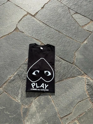 Svart Comme des Garçons Play t-shirt - Svart t-shirt från Comme des Garçons Play med det ikoniska hjärtmotivet med ögon på bröstet och vit text. Klassisk passform och rund hals. Perfekt för dig som gillar streetwear och vill sticka ut med en stilren design.