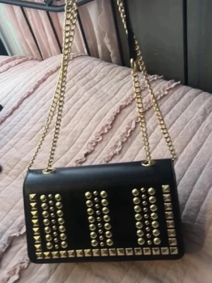 Svart axelväska med nitar och kedja - Cool svart axelväska med guldiga nitar i rader och fyrkantiga detaljer. Väskan har en lång guldfärgad kedjerem och rektangulär form. Perfekt för dig som gillar edgy stil och vill sticka ut. Materialet ser ut som konstläder.