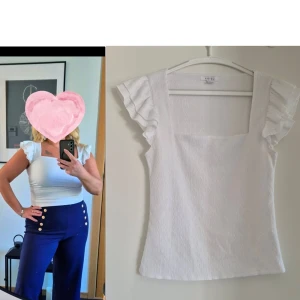 Vit blus med volangärmar - Supersöt vit blus med fyrkantig ringning och volangdetaljer på axlarna. Toppen är ärmlös med volang och har en lätt strukturerad yta. Perfekt att styla med både jeans och kjol för en trendig look.