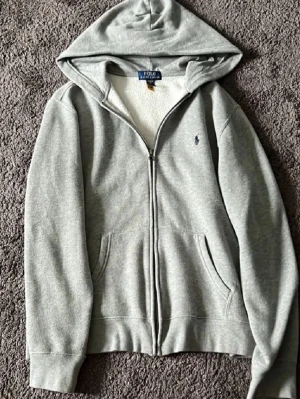 Grå zip hoodie från Polo Ralph Lauren - Snygg grå hoodie med dragkedja från Polo Ralph Lauren. Klassisk design med huva, två fickor framtill och den ikoniska lilla loggan broderad på bröstet. Tillverkad i mjuk bomullsblandning som känns skön mot huden. Perfekt för en avslappnad streetwear-look.
