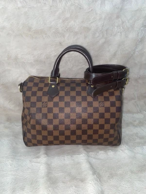 Louis Vuitton Damier Speedy handväska - Snygg handväska från Louis Vuitton i klassiskt brunt Damier-mönster. Väskan har två mörkbruna handtag, avtagbar axelrem och guldfärgade metalldetaljer. Tillverkad i canvas med läderdetaljer. Perfekt för dig som gillar lyxiga accessoarer. Det är en speedy 30 med mått 30, 21, 17 cm, inköpt år 2021 i själva Louis Vuitton butiken i Stockholm. Inga defekter alls då den bara legat i min garderob bland mina andra väskor.