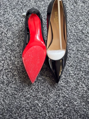 Svarta lackpumps med röd sula - Snygga svarta pumps i lack från Beikeyuan med spetsig tå och ikonisk röd sula. Skorna har en klassisk och elegant design med hög klack och beige innersula. Perfekta för dig som vill sticka ut med stil.