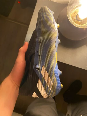 Blåa Adidas fotbollsskor med gula detaljer - Säljer ett par blåa fotbollsskor från Adidas med gula och vita detaljer. Skorna har en strömlinjeformad design utan snörning och är tillverkade i syntetmaterial. Perfekta för dig som vill ha lätta och snabba skor på planen.