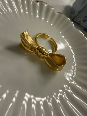 Guldfärgad ring med rosett - Säljer en chunky guldfärgad ring i metall med en stor, detaljerad rosett framtill. Ringen är öppen baktill vilket gör den justerbar i storlek. Perfekt statement piece för dig som gillar unika accessoarer och vill sticka ut. Från ”alvazessentials”💗