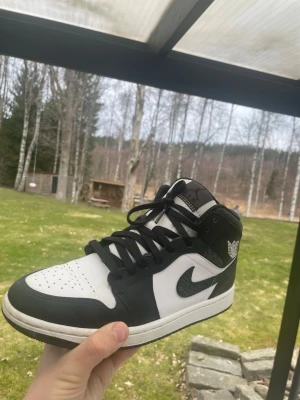 Svarta och vita Air Jordan 1 Mid - Snygga Air Jordan 1 Mid sneakers i svart och vitt läder med klassisk Swoosh-logga på sidan och Air Jordan Wings-märke vid ankeln. Skorna har svart snörning och vit tåbox med perforeringar. Perfekta för dig som gillar streetwear och ikonisk design.