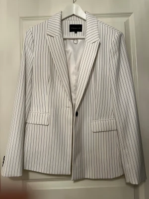 Vit randig kavaj från STOCKH LM - Snygg vit kavaj med svarta tunna ränder från STOCKH LM. Klassisk modell med slag, två fickor med lock och en knapp framtill. Fodrad insida för extra komfort. Perfekt för en stilren och trendig look.
