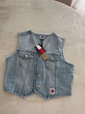 Ljusblå jeansväst från Tommy Jeans - Snygg ljusblå jeansväst från Tommy Jeans med klassisk v-ringning, tryckknappar framtill och två bröstfickor med lock. Västen har en avslappnad passform och ikonisk logga nertill. Perfekt för att lyfta din outfit med en cool denimlook.
