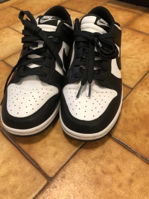 Nike Dunk Low svart/vit sneakers - Nike Dunk Low sneakers i svart och vitt läder med klassisk Swoosh-logga på sidorna. Skorna har perforerad tåbox, svart snörning och vit mellansula. Perfekt för dig som gillar en clean och tidlös stil.