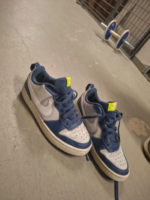 Nike blåvita sneakers med neon-detalj - Snygga sneakers från Nike i blått och vitt med klassisk swoosh på sidan. Skorna har låga skaft, perforerad tåbox och en cool neongul detalj på plösen. Ovandelen är i läder och textil, och sulan är vit med blå inslag. Perfekta för en streetwear-look.