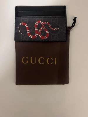 Gucci korthållare med ormmotiv - Snygg svart korthållare från Gucci med ikoniskt ormmotiv i rött, vitt och svart på framsidan. Tillverkad i canvas med läderdetaljer och diskret GG-mönster i bakgrunden. Perfekt för dig som vill ha en stilren och trendig accessoar.