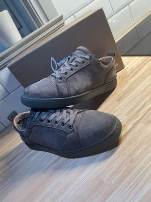 Christian Louboutins gråa med mocka - Snygga gråa sneakers från Christian Louboutin i mocka med klassisk röd sula. Lågt skaft, rund tå och matchande grå skosnören. Perfekta för dig som vill ha en clean och exklusiv look med en twist.