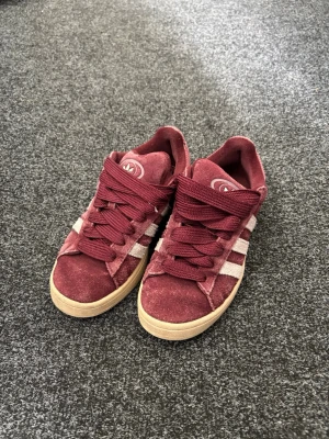 Vinröda Adidas Campus sneakers - Snygga vinröda Adidas Campus sneakers i mocka med klassiska vita ränder på sidorna och beige sula. Skorna har lågt skaft och breda vinröda skosnören. Perfekta för dig som gillar retrostil och streetwear.