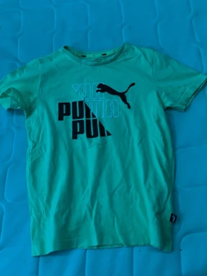 Puma t-shirt med logga - Snygg grön t-shirt från Puma med stor svart logga och text på bröstet. Klassisk rund halsringning och korta ärmar. Tillverkad i mjuk bomull som känns skön mot huden. Perfekt för dig som gillar sportig och avslappnad stil.