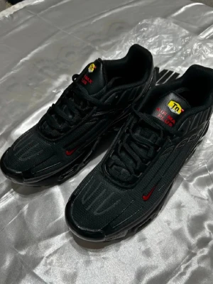 Nike Air Max Plus svarta sneakers - Nike Air Max Plus sneakers i helsvart med röda detaljer och Tn-logga i gult på plösen. Skorna har en sportig design med synliga Air Max-enheter i sulan och diskreta mönster på ovandelen. Perfekta för dig som gillar streetwear och vill sticka ut med stil.