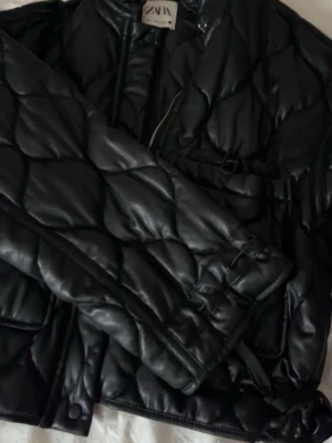 Svart pufferjacka från Zara - Snygg svart pufferjacka från Zara med quiltat mönster och glansig finish. Jackan har hög krage, dragkedja framtill och bälte i midjan för en schysst siluett. Perfekt för kalla dagar när du vill hålla stilen.