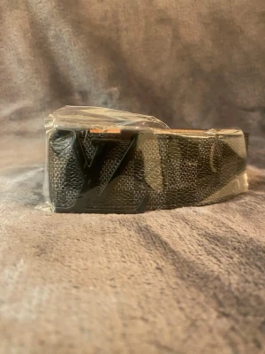 Svart Ioois Vuiton bälte med Iv-spänne - Snyggt svart bälte från Iv med det ikoniska Iv-spännet i svart metall. Bältet har diskret mönster på bandet och insidan är beige. Perfekt accessoar för att lyfta din outfit.