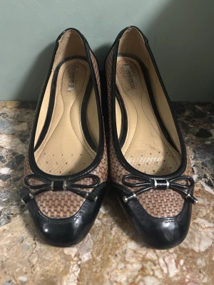 Bruna ballerinaskor med flätad design - Snygga ballerinaskor från Geox i brun flätad textil med svarta lackdetaljer och en liten rosett framtill. 