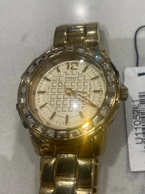 Guldig klocka från Guess med stenar - Snygg klocka från Guess i guldigt metallutförande med glittriga stenar runt urtavlan. Urtavlan har ett geometriskt mönster och tydliga markeringar. Länken är bred och ger ett lyxigt intryck. Perfekt accessoar för att lyfta din stil.
