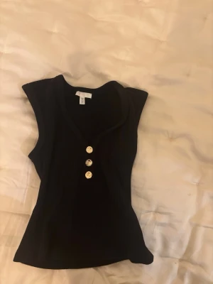 Svart ribbad ärmlös topp med knappar - Snygg svart ribbad topp från H&M med v-ringning och guldfärgade knappar framtill. Toppen är ärmlös och har en figurnära passform som framhäver siluetten. Kan postas idag🩵