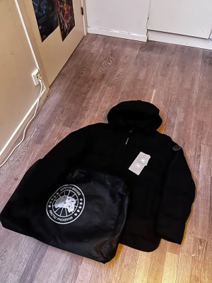 Svart dunjacka från Canada Goose - Snygg svart dunjacka från Canada Goose med huva och diskret logga på ärmen. Jackan har dragkedja framtill och en klassisk puffad design som håller dig varm under kalla dagar. Medföljer originalpåse med Canada Goose Arctic Program-tryck.