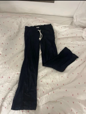 Mörkblå linnebyxor från Roxy - Snygga mörkblå linnebyxor från Roxy med avslappnad passform och resår i midjan. Byxorna har en vit dragsko framtill och raka ben. Perfekta för en chill och bekväm stil. Materialet är lätt och luftigt linne. Byxorna är svarta/mörkblå