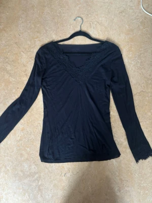 Svart långärmad topp med spets från Intimissimi  - Svart långärmad topp med v-ringning och dekorativ spets runt halsen och vid ärmsluten. Tunn och mjuk i materialet, perfekt för lager-på-lager. Enkel och stilren design som funkar till många olika looks. Finns ett litet hål på ryggen men det syns knappt. 