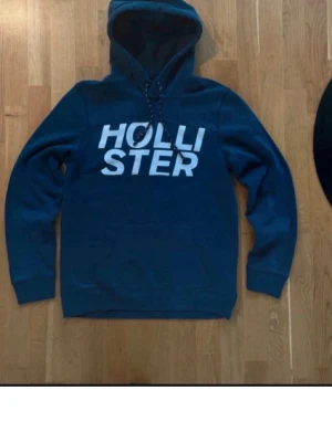 Mörkblå hoodie från Hollister - Snygg mörkblå hoodie från Hollister med stor vit logga på bröstet. Klassisk huva med dragsko och magficka framtill. Tillverkad i mjukt material som känns skönt mot huden. Perfekt för en avslappnad och trendig look.