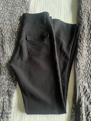 Svarta kostymbyxor med ficka - Snygga svarta kostymbyxor med rak passform och en bakficka med knapp. Byxorna har hällor för skärp och stilren design. Perfekta för en clean och dressad look. Materialet ser ut att vara en mjuk och stretchig blandning, vilket ger skön komfort.