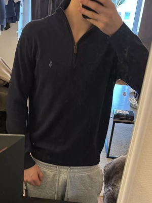 Mörkblå halvzip från Polo Ralph Lauren - Mörkblå tröja från Polo Ralph Lauren med halv dragkedja och ståkrage. Klassisk broderad logga på bröstet och ribbade detaljer vid kragen. Perfekt för en stilren och avslappnad look.