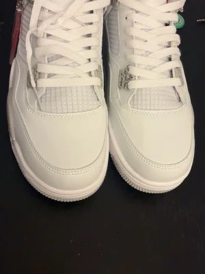 Vita Air Jordan 4 Retro sneakers - Snygga vita Air Jordan 4 Retro sneakers med detaljer i grått och svart. Skorna har meshpaneler, klassiska plastvingar och synlig Air-sula. Ovandelen är i läder och textil, och sulan har stänkdetaljer. Perfekta för dig som gillar streetwear och ikoniska sneakers.