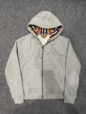 Grå zip hoodie från Burberry - Säljer en grå zip hoodie från Burberry med klassiskt rutigt foder i huvan och broderad logga på bröstet. Hoodien har dragsko i huvan, två fickor fram och är tillverkad i mjuk bomullsblandning. Perfekt för en avslappnad och stilren look.